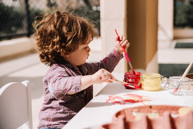 Toddler seated at table outdoors painting with primary colours