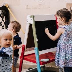 Preschooler drawing on easel with two toddlers close by