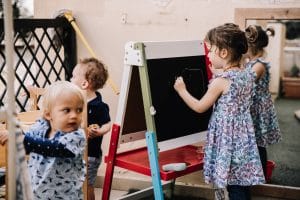 Preschooler drawing on easel with two toddlers close by